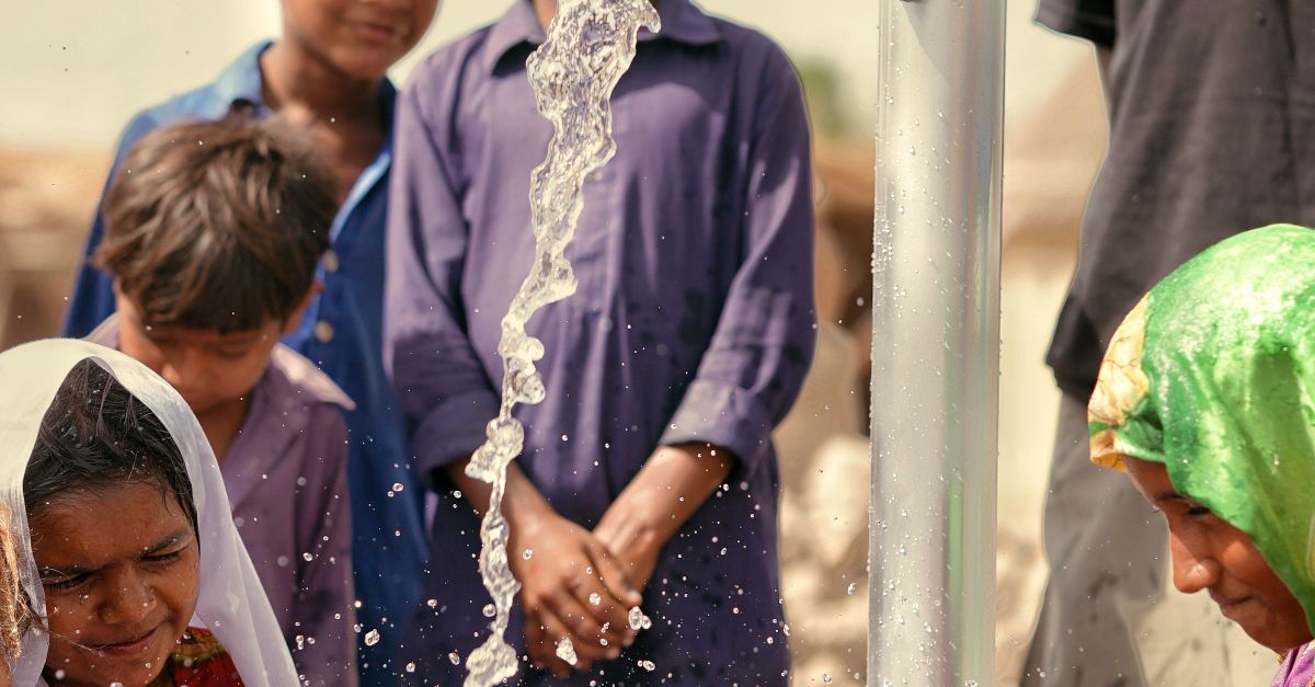 Children and women in traditional clothing using a hand pump for water supply outdoors.