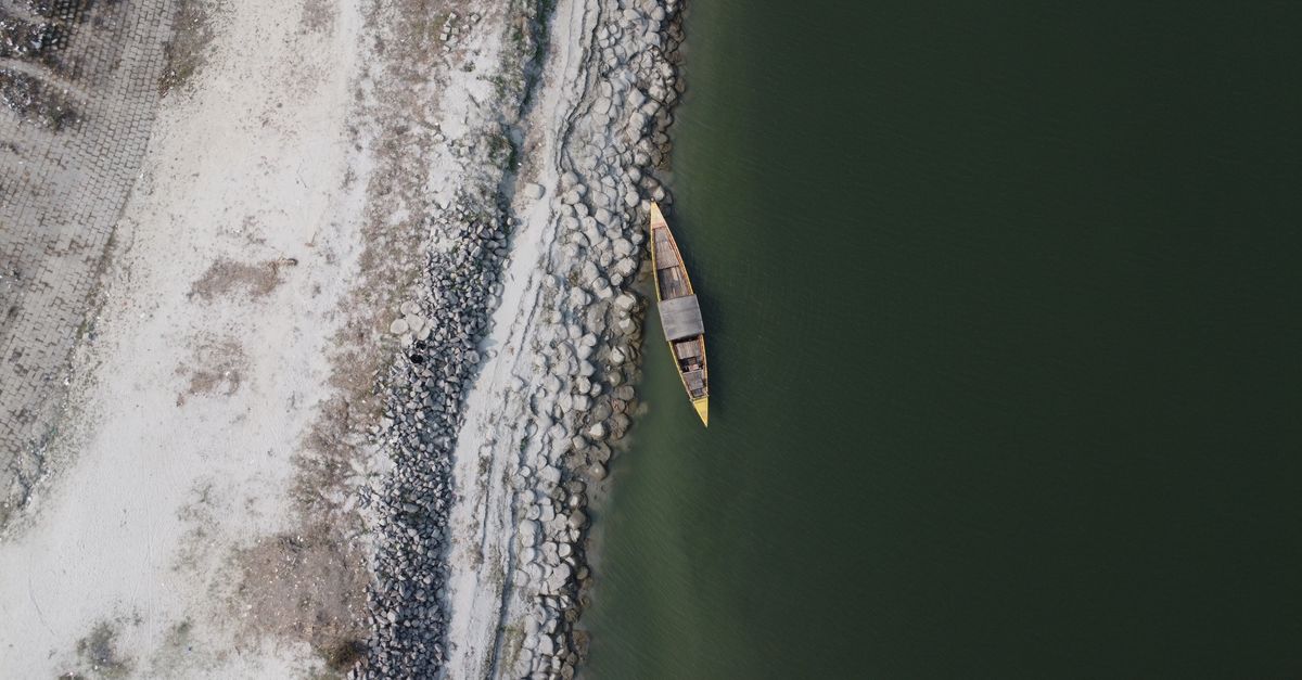 Aerial shot of a solitary wooden boat on a rocky riverbank with green water.