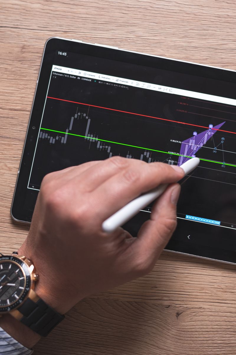 Close-up of a hand using a stylus on a digital trading app on a tablet indoors.